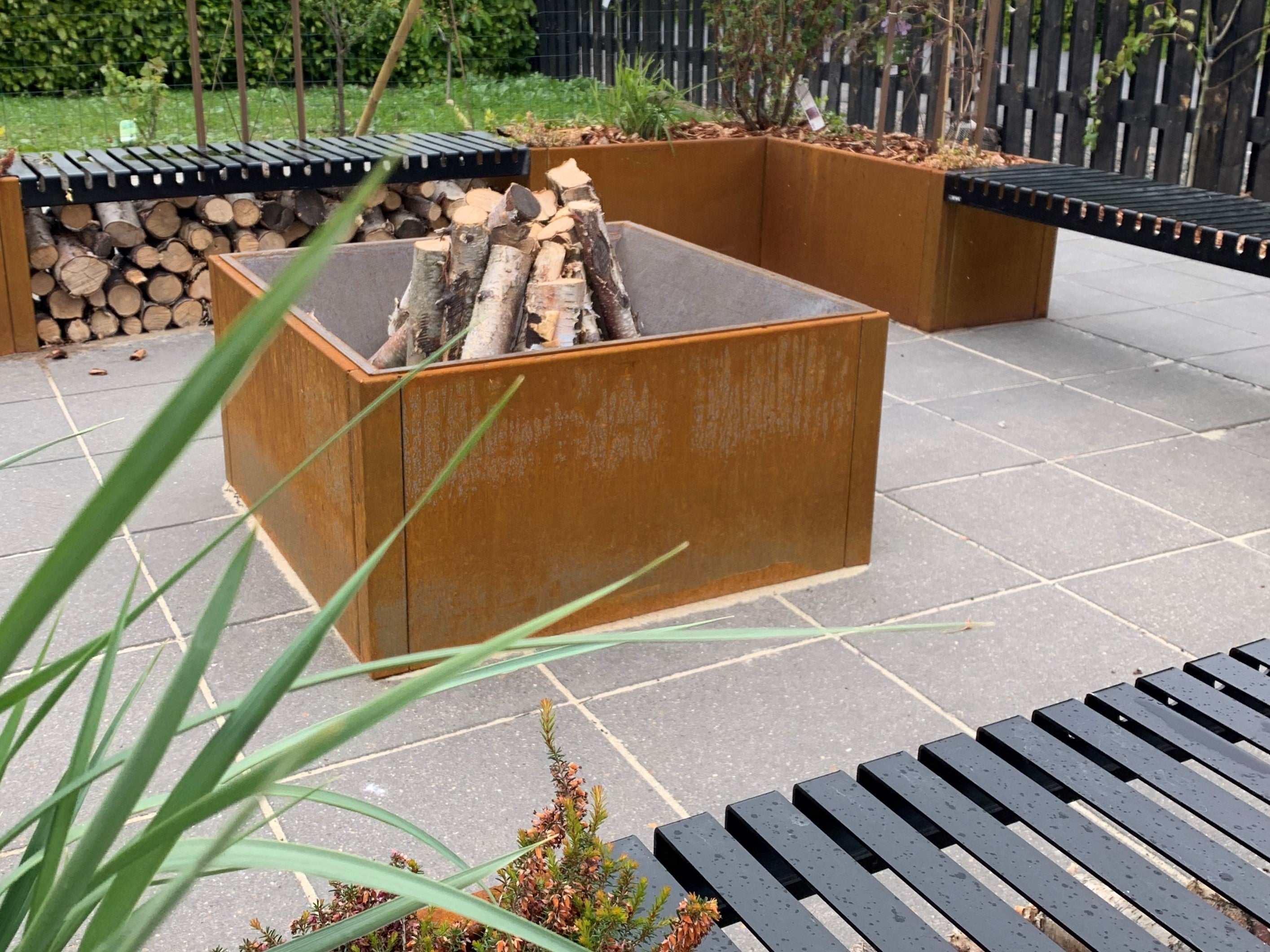 Quadratische Feuerschale aus Cortenstahl mit gestapeltem Brennholz, integriert in eine moderne Gartenterrasse mit Sitzbänken.