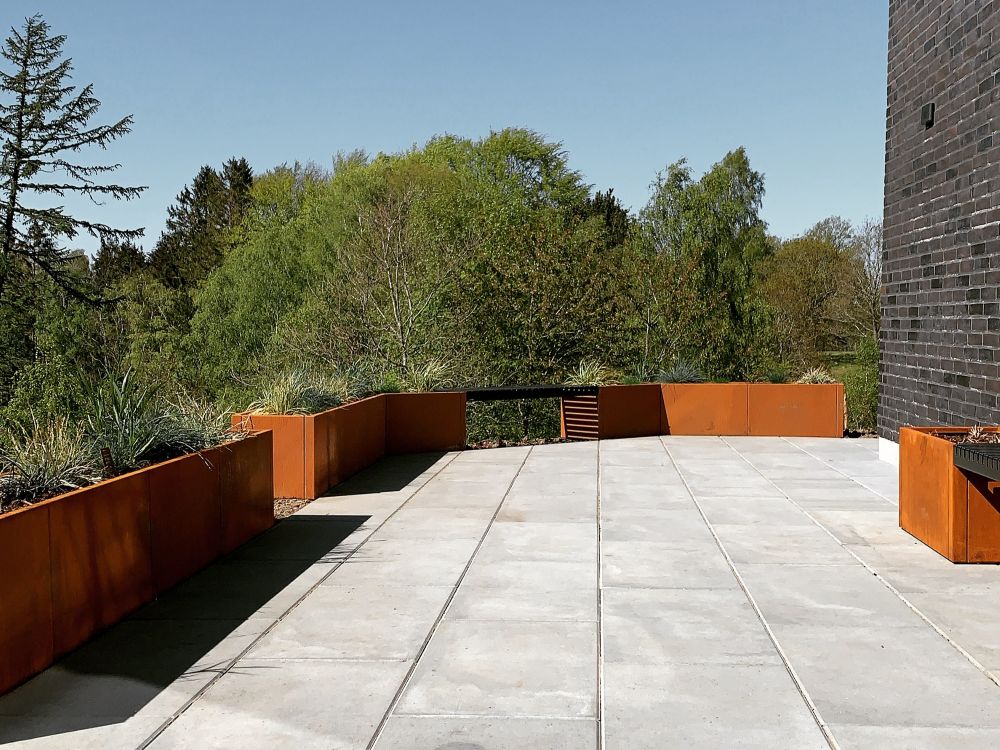 Reihe von byJEMA Pflanzkübeln aus Cortenstahl entlang einer Terrasse im Garten