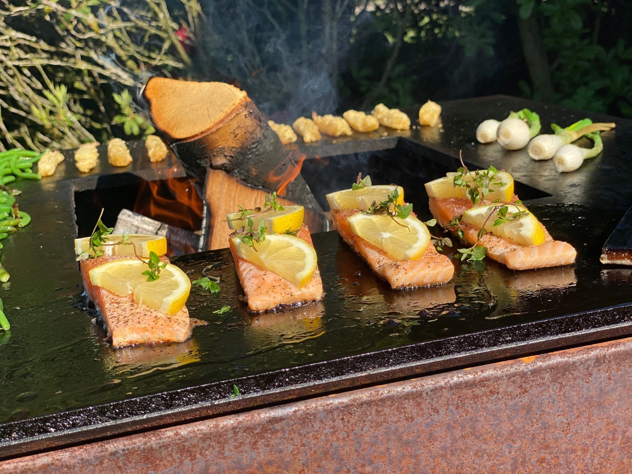 byJEMA Feuerplatte aus Cortenstahl mit offenem Holzfeuer – Lachsfilets mit Zitronenscheiben und frischen Kräutern beim Grillen im Garten